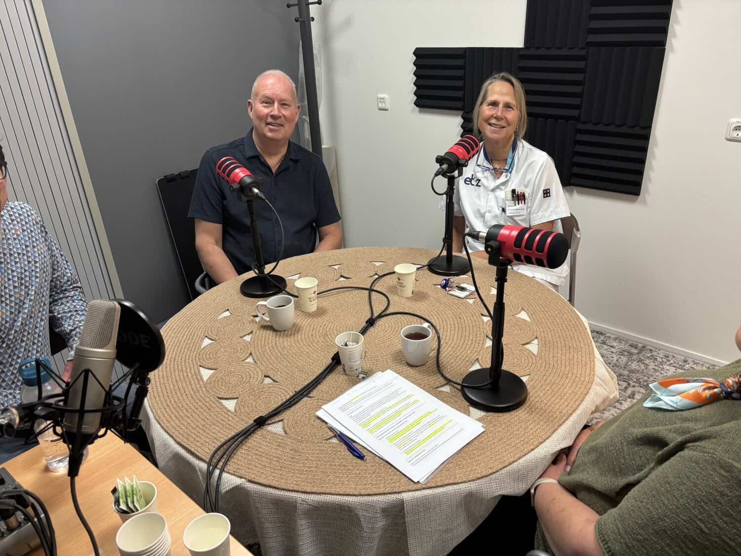 Achter een tafel met rode microfoons nemen mensen een podcast op.
